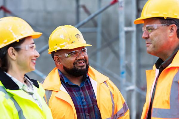 three construction workers in yellow hard hats and jackets stand together smiling and talking